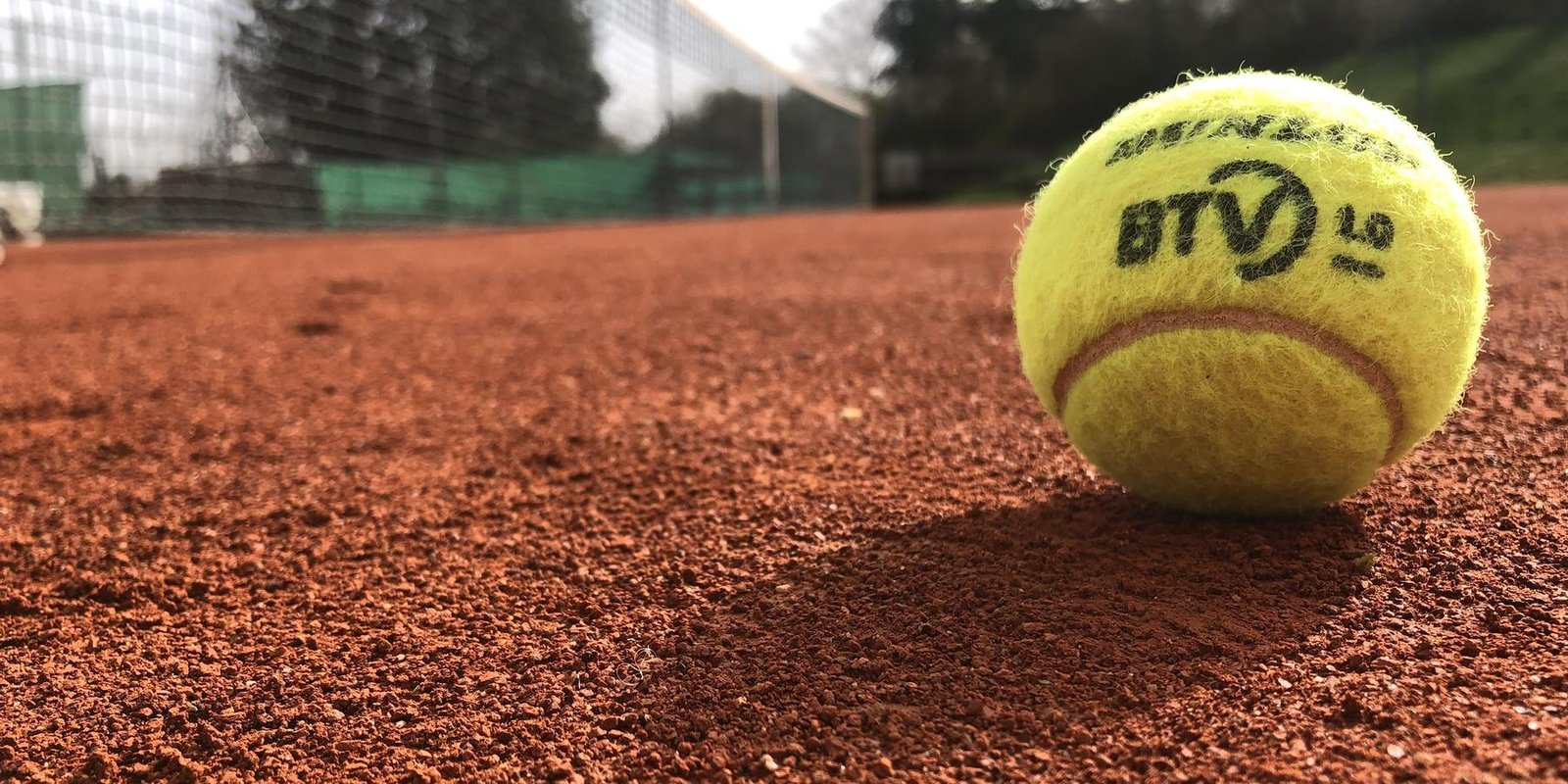 Ein Tennisball auf rotem Sand, im Hintergrund das Netz